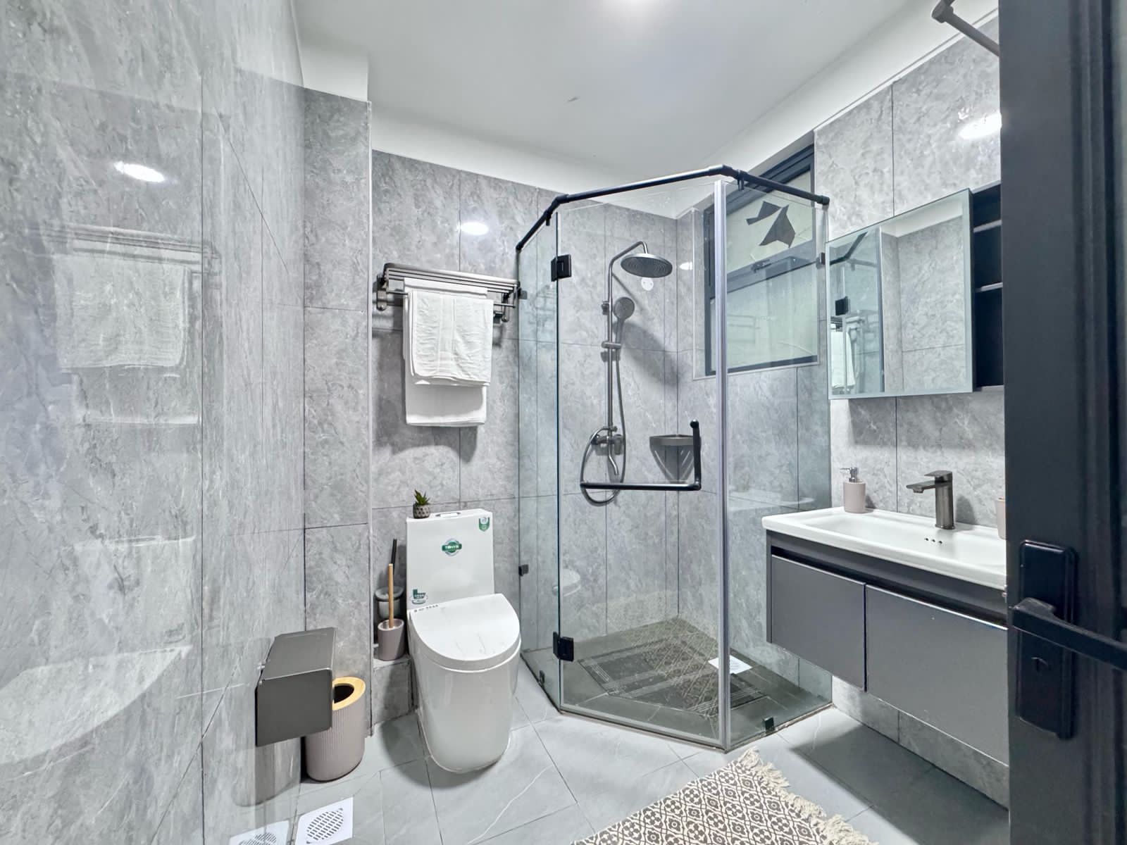 Kilimani apartment bathroom with grey wall tiles, glass-enclosed shower, toilet and wall-mounted vanity beneath a mirror.