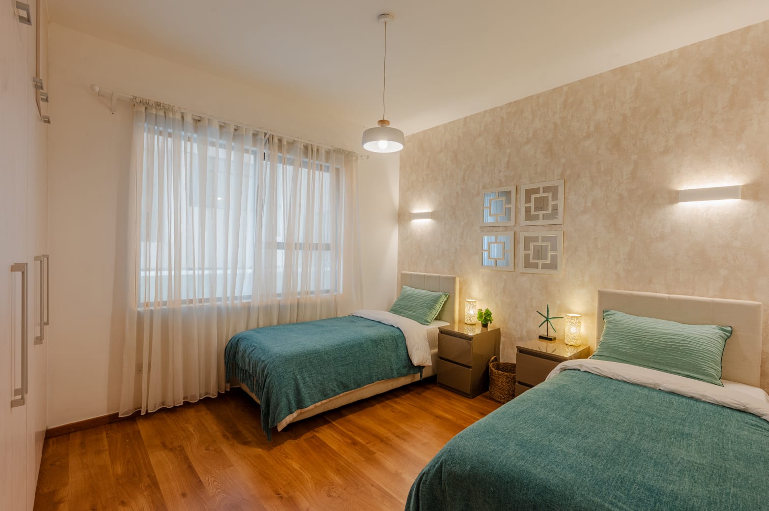 Kilimani apartment bedroom with two beds, teal bedding, bedside lamps and sheer curtains over a large window.