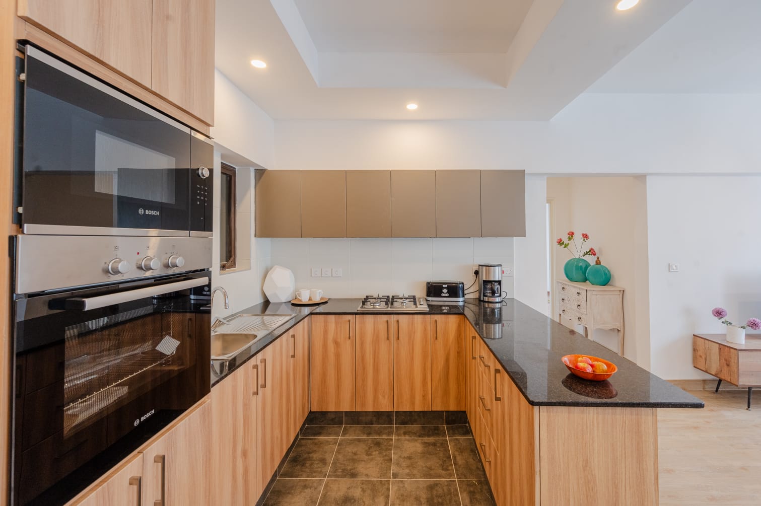 Kilimani apartment kitchen with wood-look cabinetry, black countertops, built-in oven and microwave, and a gas hob.