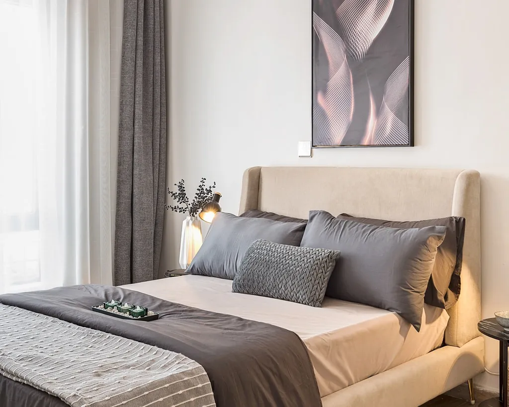 Bedroom in the South C apartment with a beige upholstered bed, grey bedding and framed abstract artwork.