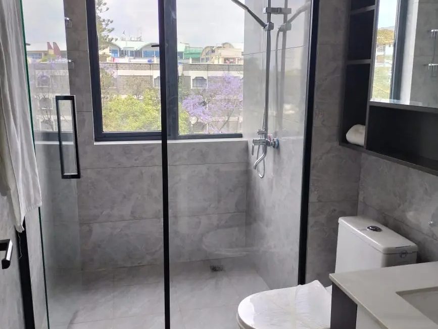Bathroom at Versailles Garden, Kileleshwa, with grey tiles, a glass shower enclosure, large window and toilet.
